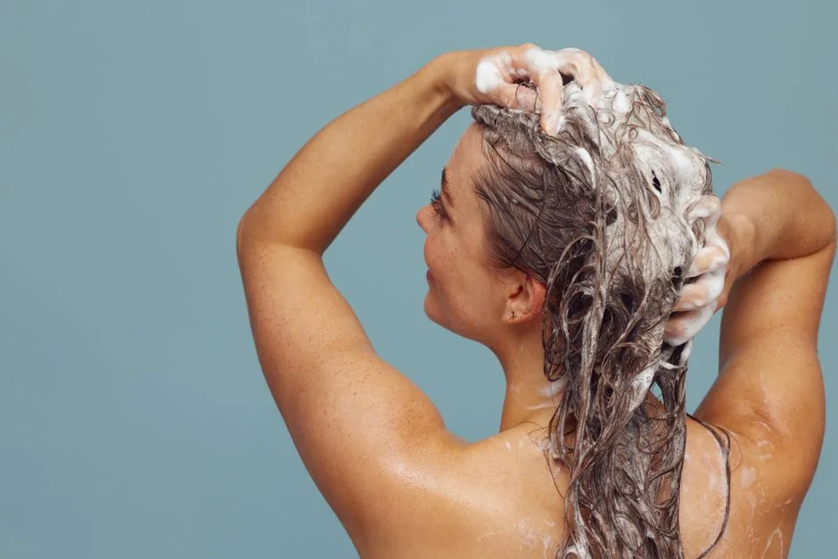 Woman with long hair washing it with shampoo, creating lather, in indoor setting against blue background.