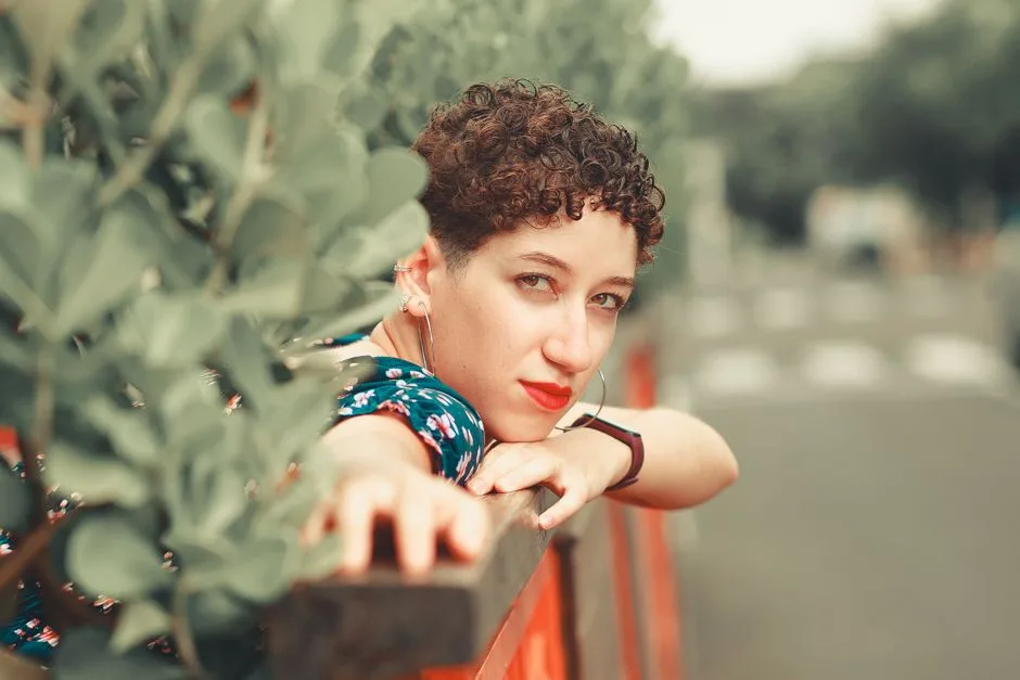 Stylish woman with curly hair leaning against a railing outdoors, wearing a smartwatch.